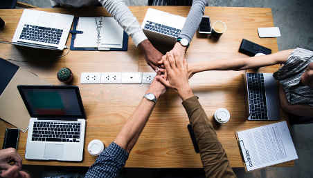 Un grupo de personas entrelazan sus manos sobre una mesa de trabajo.