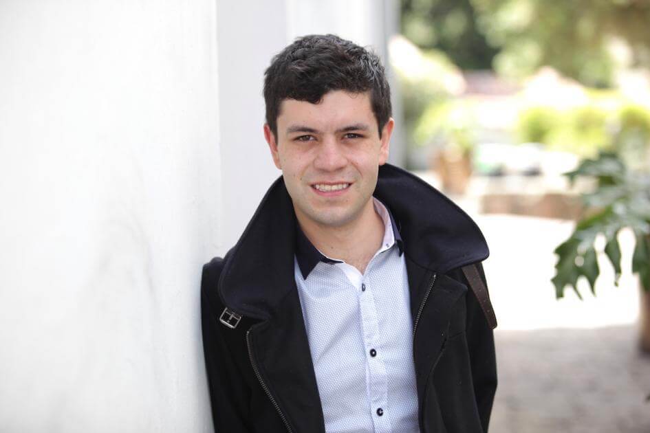 estudiante recostado sobre pared. Usa chaqueta negra y camisa blanca