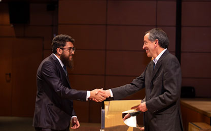 Hombre de barba recibiendo diploma y saludando a quien le hace entrega de su título.