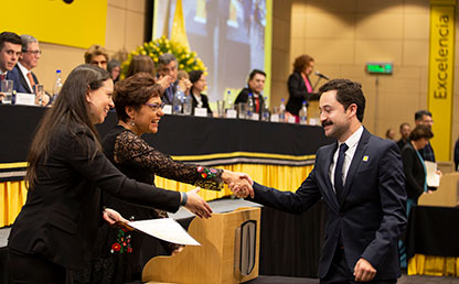 Graduando recibiendo su diploma por parte de dos mujeres en auditorio.