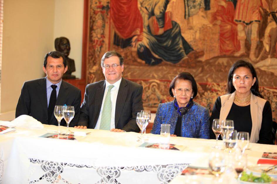 cuatro personas sentadas en una mesa, los hombres visten traje con saco y corbata, las mujeres tienen vestido
