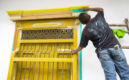 Hombre pinta una ventana de una casa de color amarillo. 